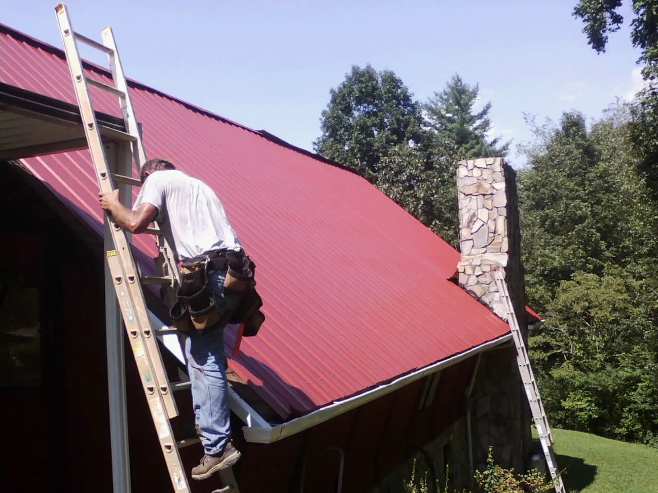 Roofer on a ladder beside a red metal roof and stone chimney — Soffit & Fascia Repair by Roofing Brothers in Richmond Hill